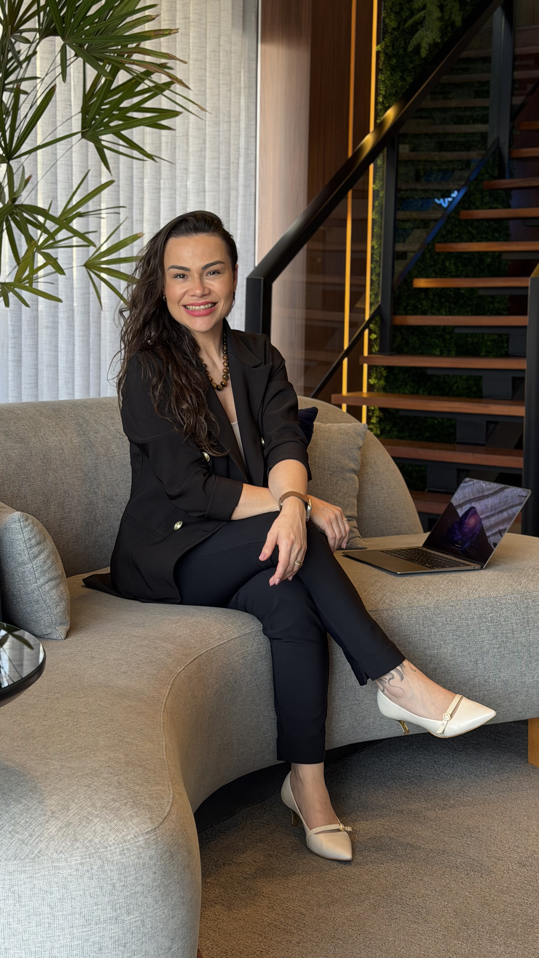 Retrato de mulher sorridente, cabelo longo e escuro, vestindo blazer preto, sentada em um sofá cinza claro em um ambiente moderno e sofisticado com escada de madeira e parede verde ao fundo, luz natural suave, fotografia profissional