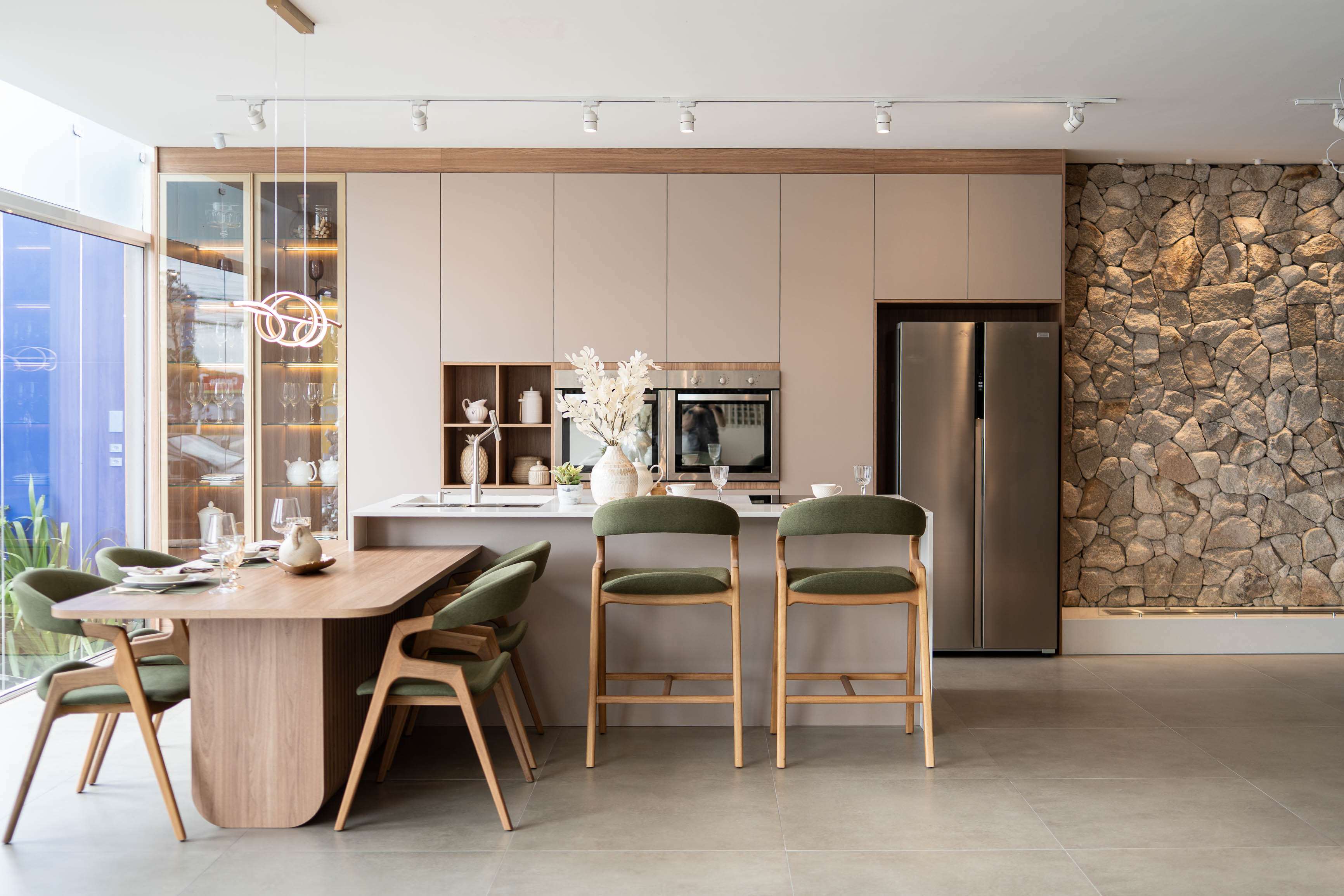 Cozinha planejada em tons terrosos integrada à sala de jantar, mesa de madeira com cadeiras estofadas em verde-musgo, cristaleira iluminada, pendente em neon dourado, parede de pedra natural ao fundo, geladeira inox, piso porcelanato cinza, fotografia de interiores realista