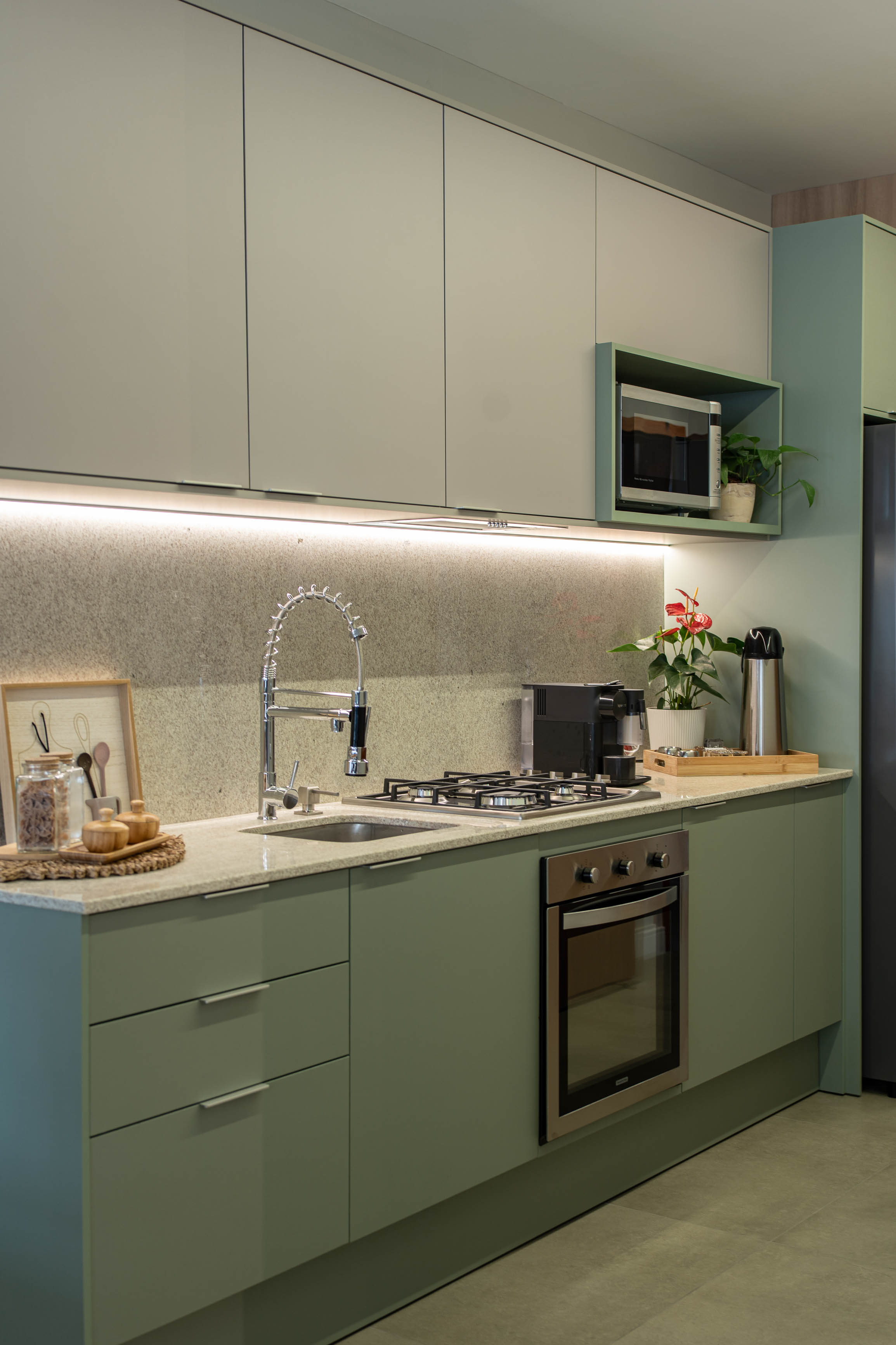 Cozinha planejada verde-sálvia e branca com bancada em quartzo branco, nicho iluminado com micro-ondas, coifa em inox, torneira gourmet cromada, cafeteira e vasos com flores, piso porcelanato claro, fotografia de interiores realista