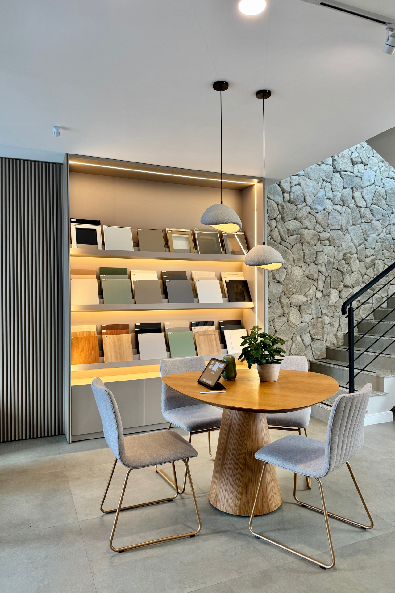 Sala de estar sofisticada com mesa redonda de madeira sobre pedestal, cadeiras estofadas cinza com pés dourados, estante de amostras de revestimentos e acabamentos iluminada em LED, parede de pedra natural ao fundo, escada com guarda-corpo em cabo de aço, vaso de planta sobre a mesa, iluminação quente aconchegante, fotografia de interiores realista, showroom
