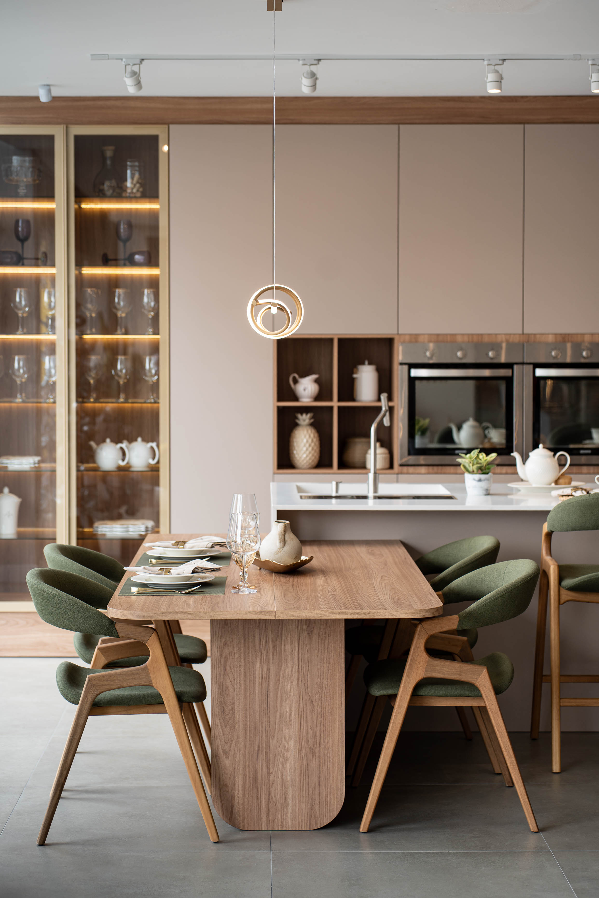 Sala de jantar planejada integrada à cozinha, mesa de madeira com cadeiras estofadas em verde-musgo, cristaleira com iluminação interna e taças de cristal, pendente dourado circular, bancada em quartzo branco ao fundo, fotografia de interiores realista