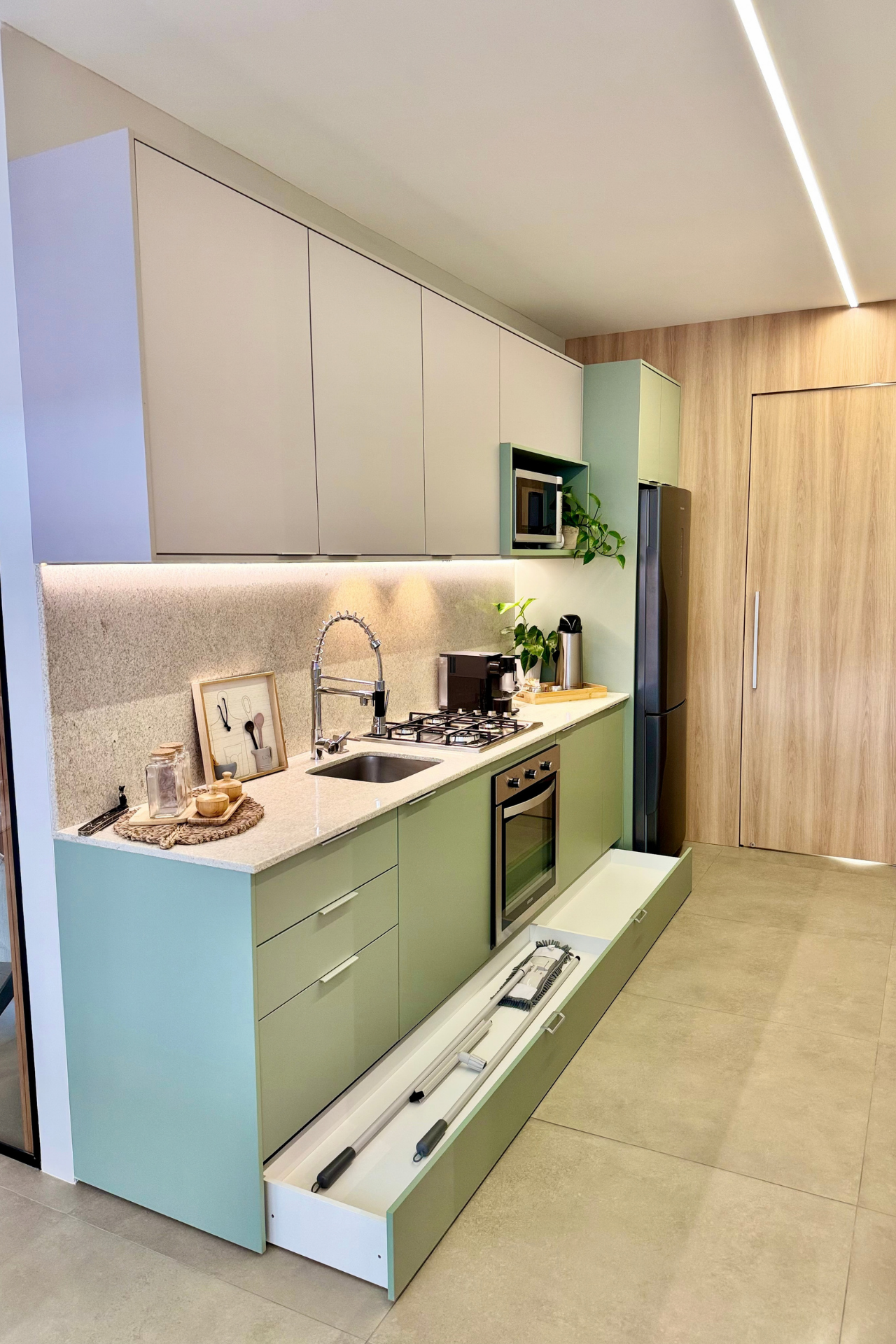 Cozinha planejada bege e verde-sálvia com bancada em granito claro, coifa e cooktop embutidos, armário superior suspenso, micro-ondas de embutir, geladeira inox, porta de madeira amendoada ao fundo com iluminação em friso de LED, fotografia de interiores realista