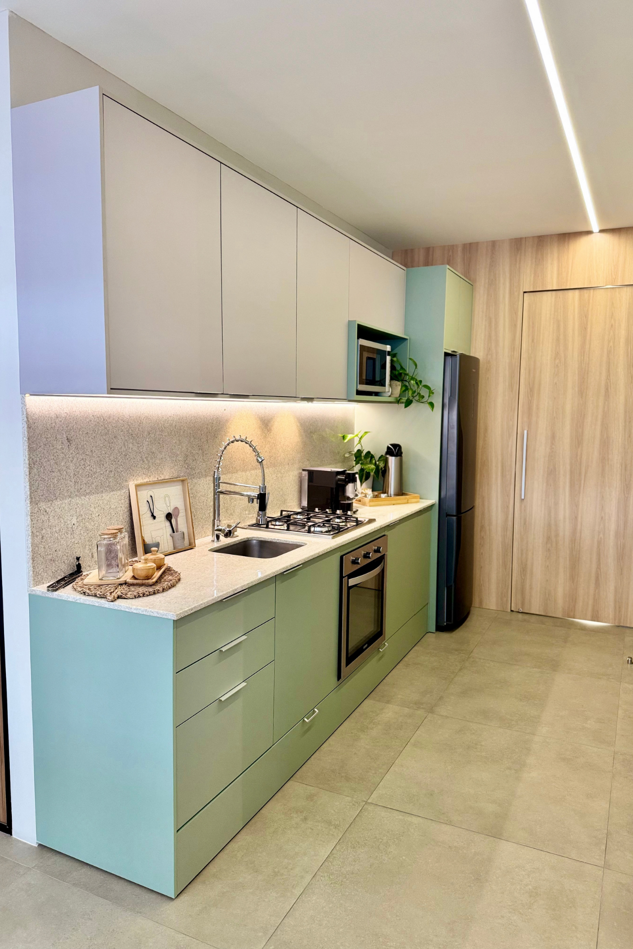 Cozinha planejada verde-sálvia vista em perspectiva com bancada em granito claro, coifa e cooktop embutidos, armário suspenso bege, geladeira inox, porta de madeira amendoada ao fundo com iluminação em friso de LED, piso porcelanato cinza, fotografia de interiores realista
